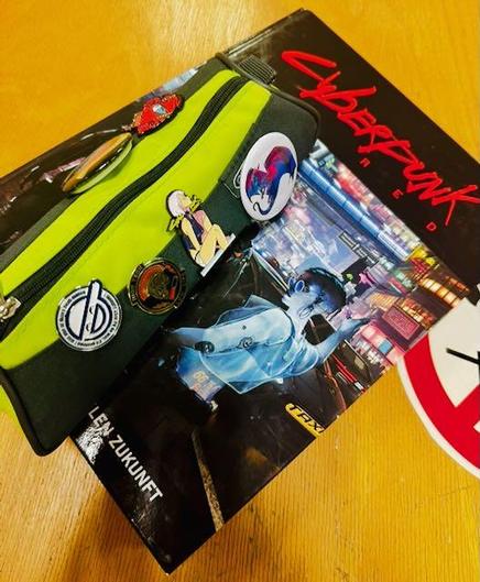 Auf einem abgenutzten Holztisch liegt das Cyberpunk RED Rollenspielregelbuch. Darauf liegt ein Federmäppchen mit diversen Pins, u.a. der Smiling Man aus GITS.