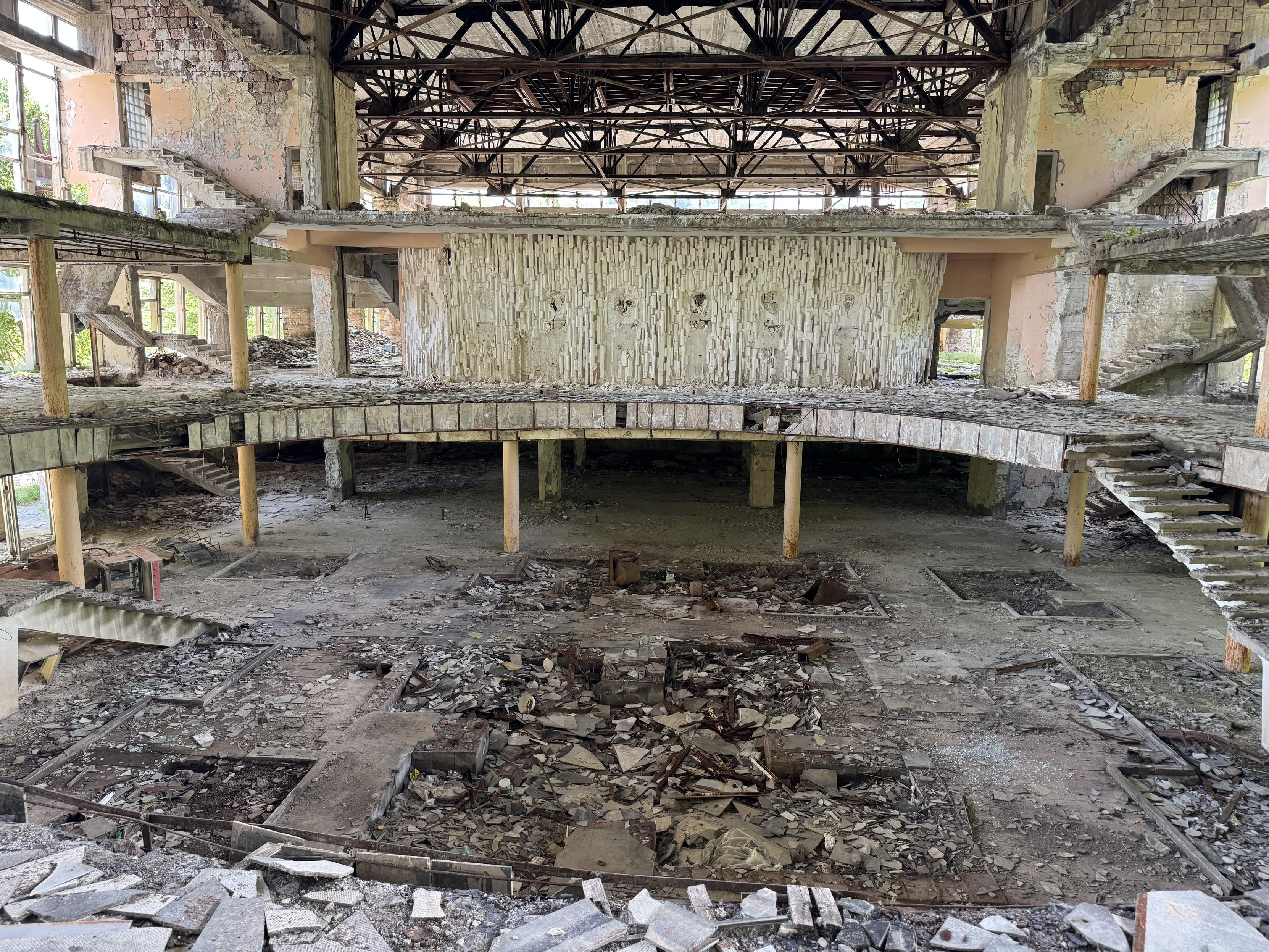 Innenraum eines verfallenden Gebäude. Große Halle mit mehreren Ebenen, im Zentrum eine große Halle. Auf den Böden Schutt, von den Treppen nur noch das Beton-Skelett vorhanden. An der Decke die blanke Stahlkonstruktion.