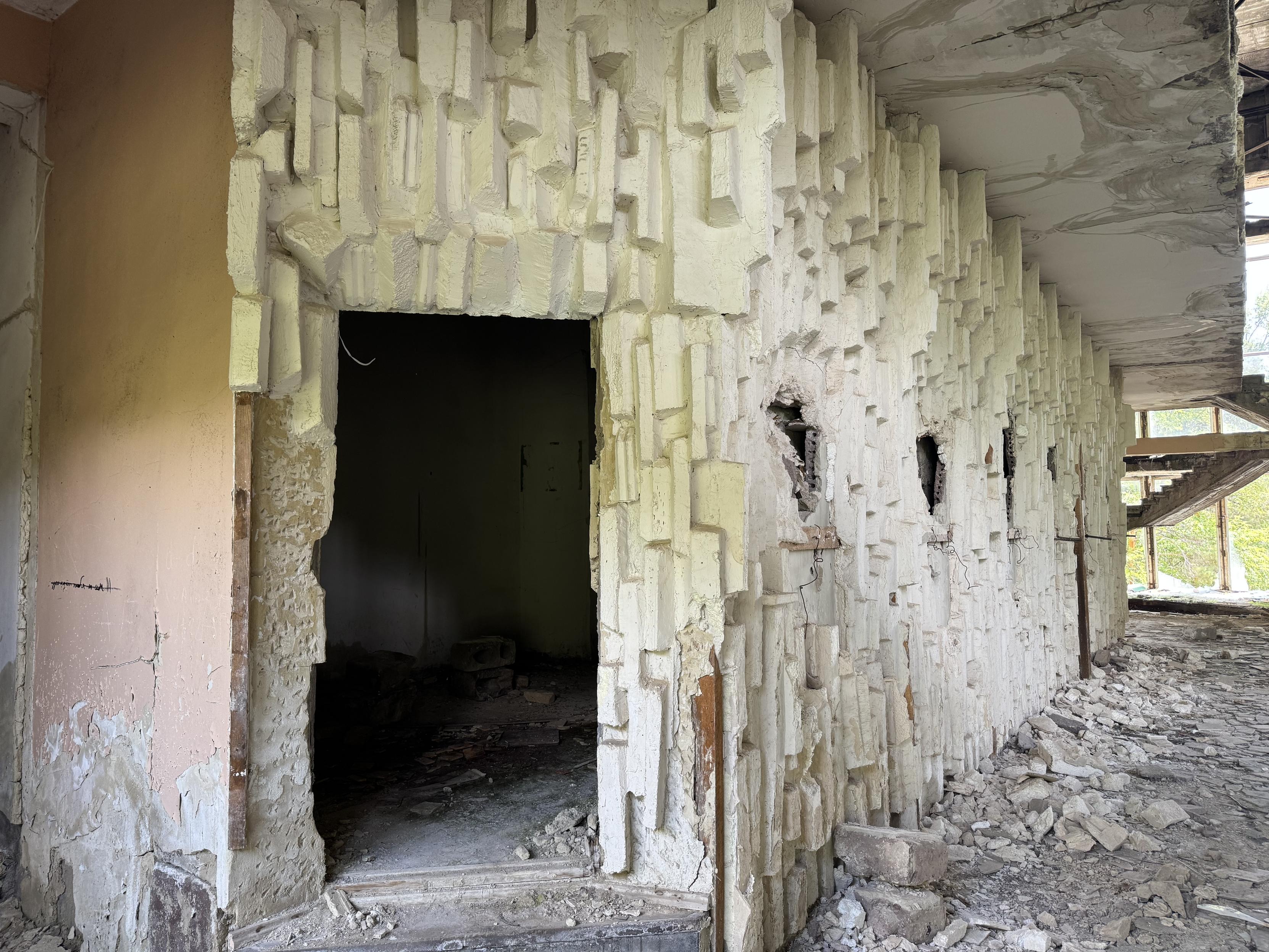 Eine Wand mit unregelmäßiger Struktur. Im Zentrum eine Tür, die in einen dunklen Raum führt. In der Wand einige Löcher. Am Boden Schutt.